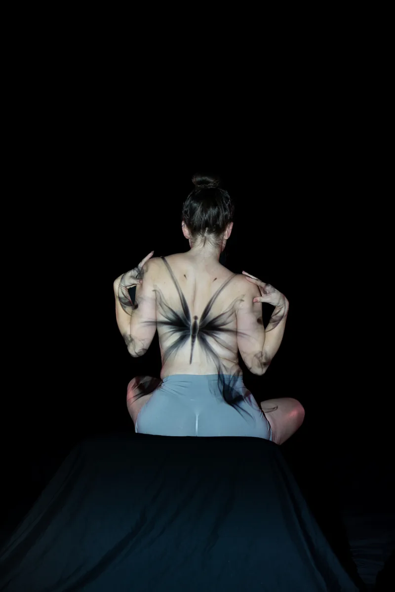 A person with hair in a bun sits facing away, with a dark butterfly and abstract shapes projected onto their bare back and arms against a black background.